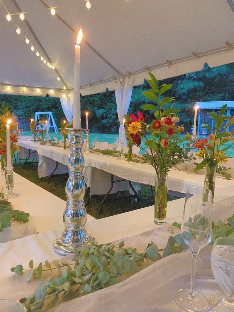 photo of white long tables under a white tent with hanging lights and tall bud vases on the table