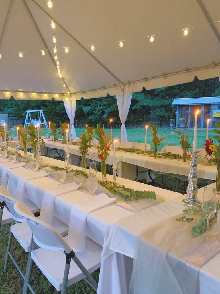 photo of white long tables under a white tent with hanging lights and tall bud vases on the table