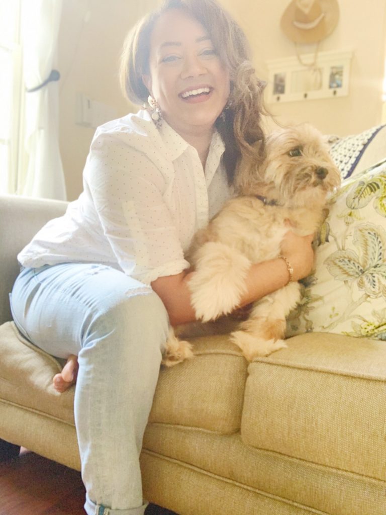 woman wearing a white button up shirt and jeans smiling at camera holding her small tan fluffy dog